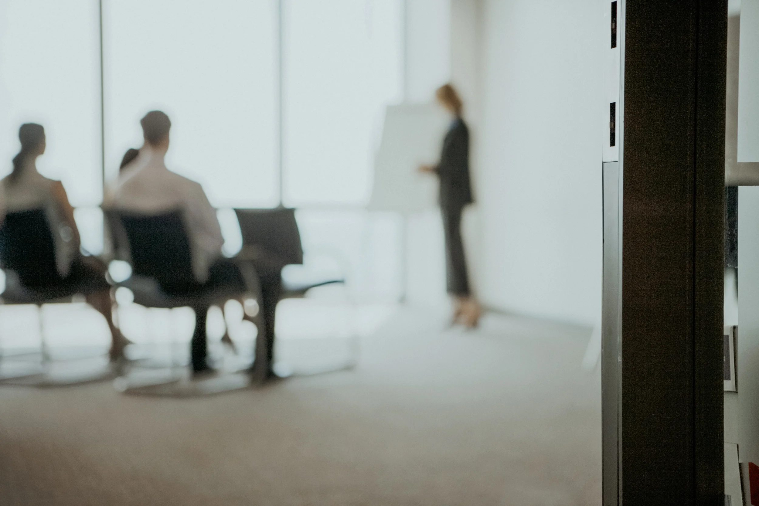 Verschwommener Blick in einen Sitzungsraum, in dem eine Frau am Flipchart vor sitzenden Kolleginnen und Kollegen präsentiert